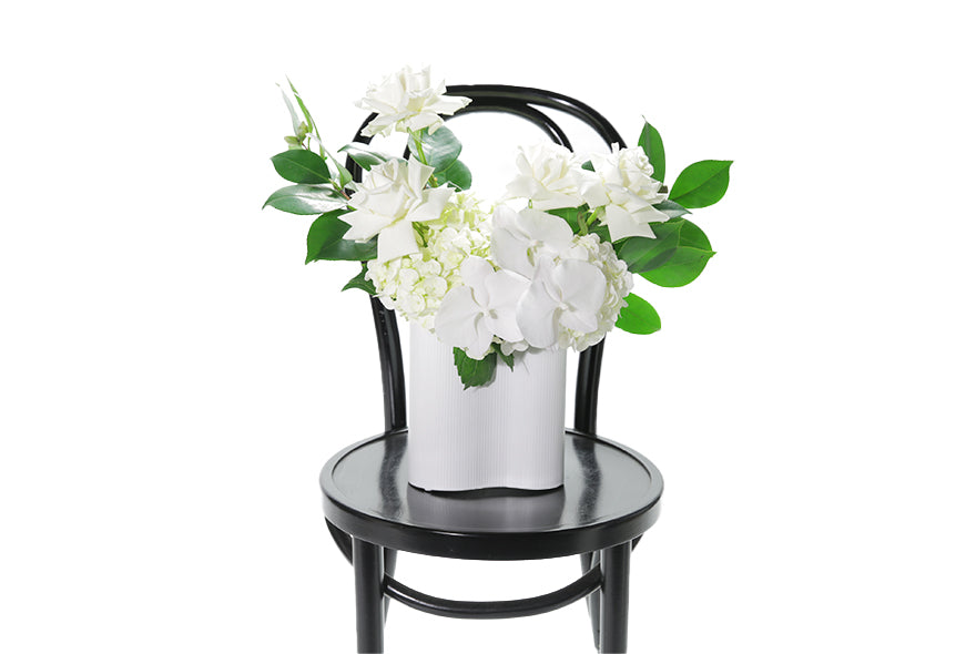 White ceramic wave vase displaying white and green seasonal flowers and foliages. Sia wave vase sitting on black bentwood chair with white background.