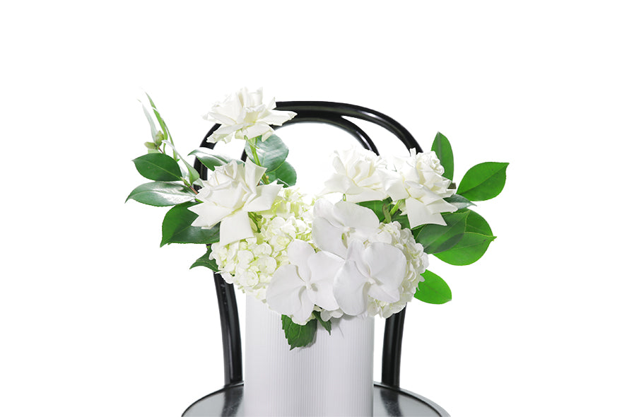 White ceramic wave vase displaying white and green seasonal flowers and foliages. Sia wave vase sitting on black bentwood chair with white background. Close up image of the flowers on black bentwood chair.