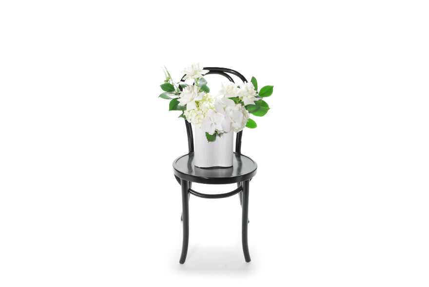 White ceramic wave vase displaying white and green seasonal flowers and foliages. Sia wave vase sitting on black bentwood chair with white background. Wide image of the white wave vase design on chair.