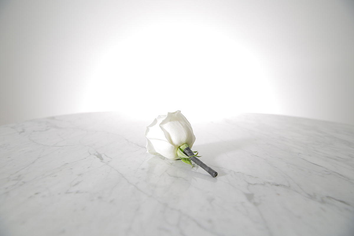 Kate Hill Flowers white rose buttonhole flower with black-taped stem and pearl pin, lapel-ready on a marble surface with a light grey background.