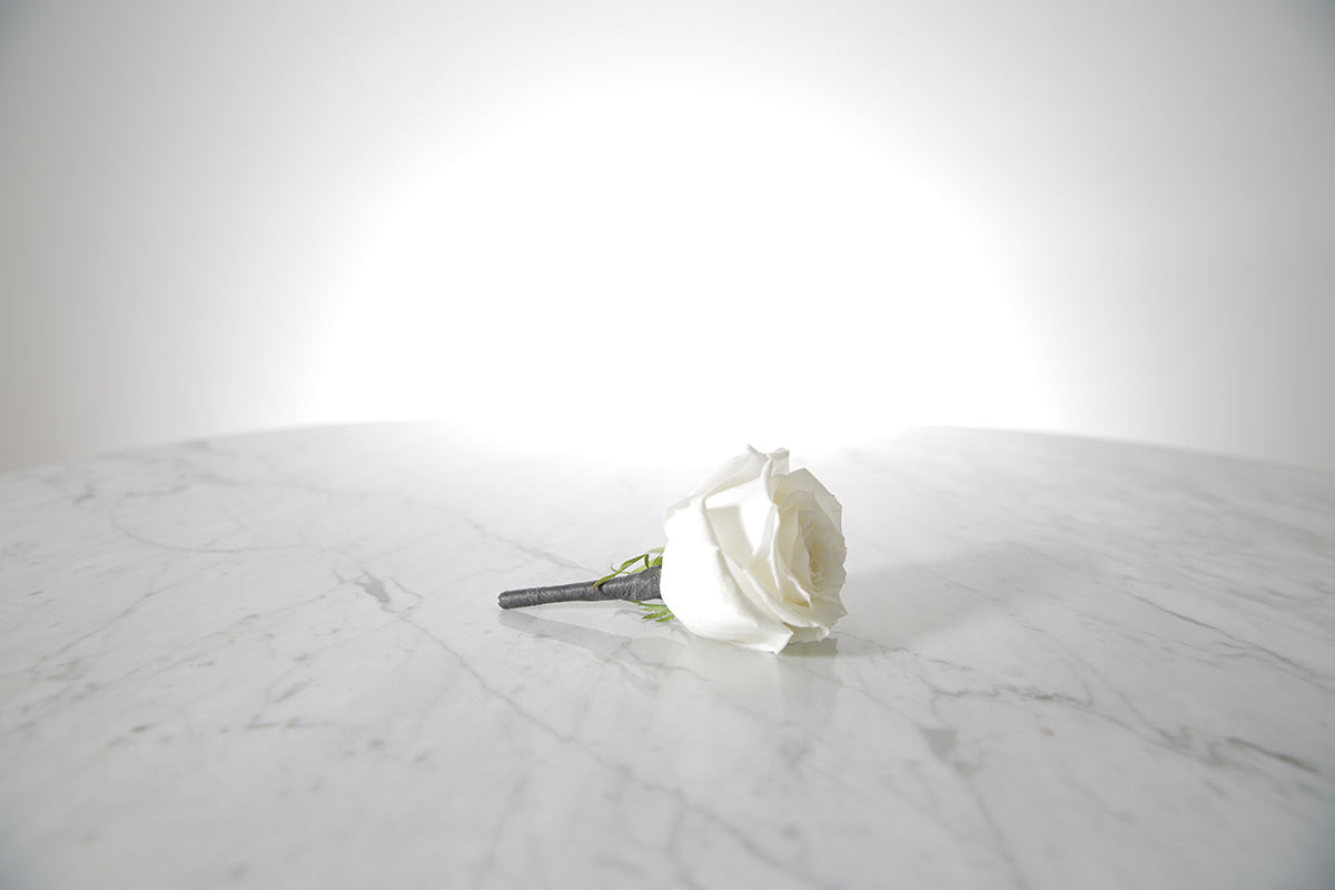 Kate Hill Flowers white rose buttonhole flower with black-taped stem and pearl pin, lapel-ready on a marble surface with a light grey background.