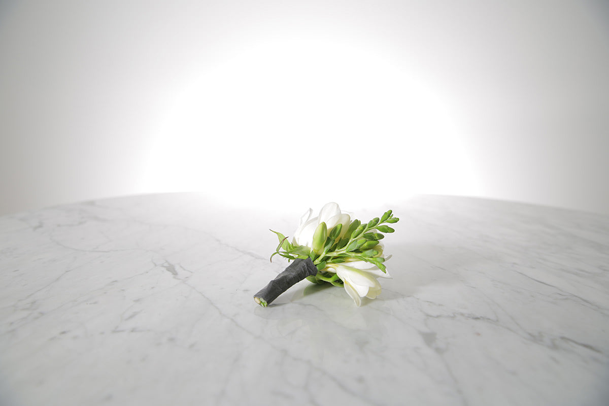 White rose and freesia buttonhole with black-taped stem and pearl pin with a light grey background