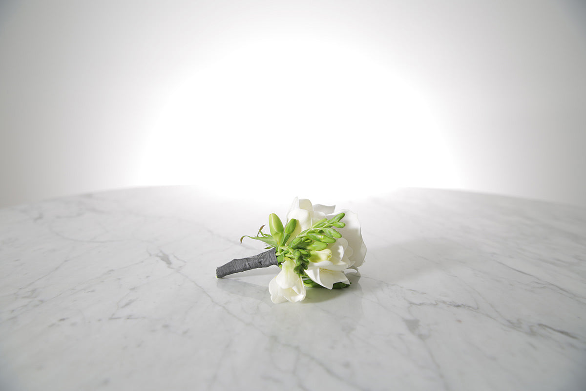 White rose and freesia buttonhole with black-taped stem and pearl pin with a light grey background