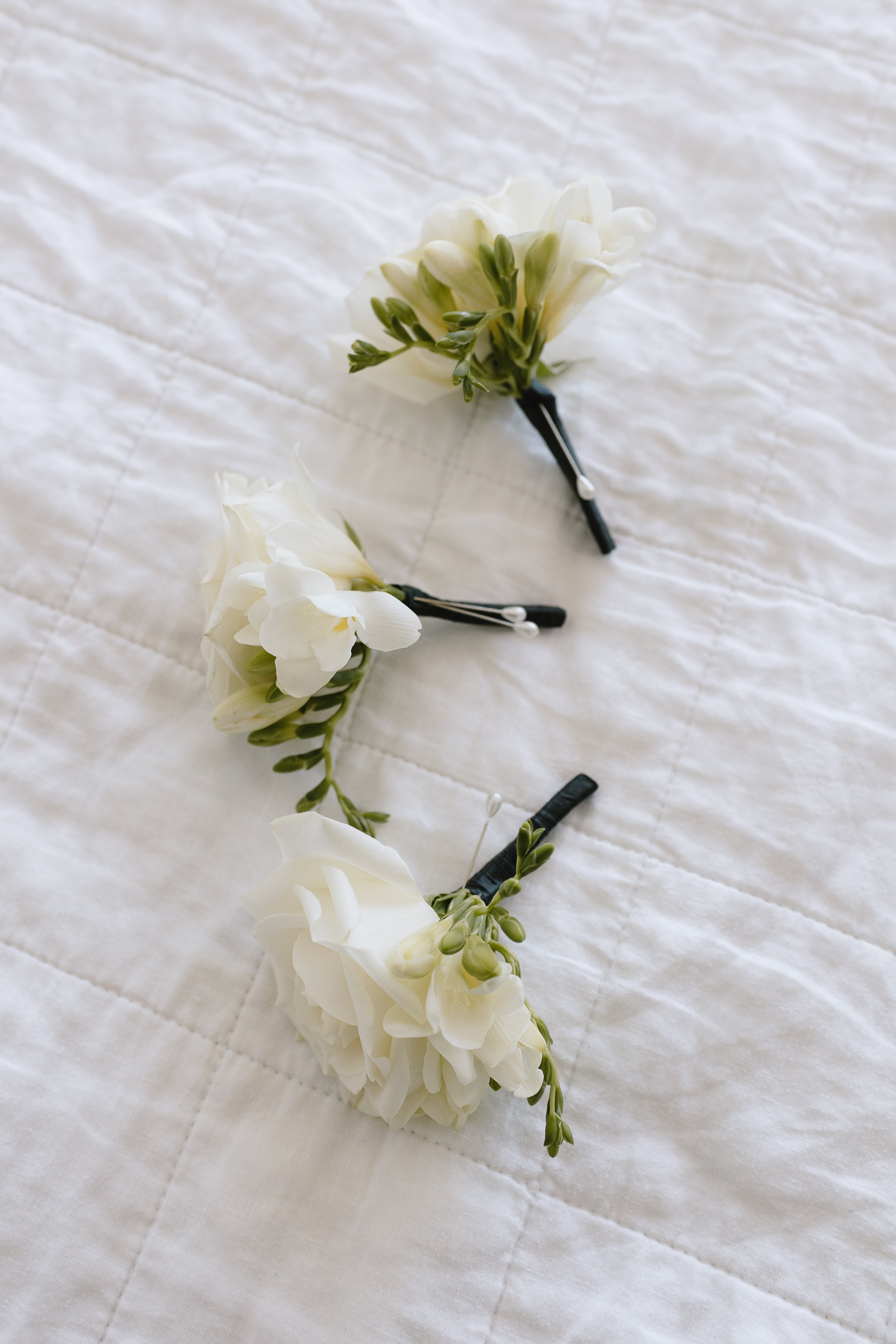 3 Buttonholes made with white freesias and black stems, laying on white linen bed.