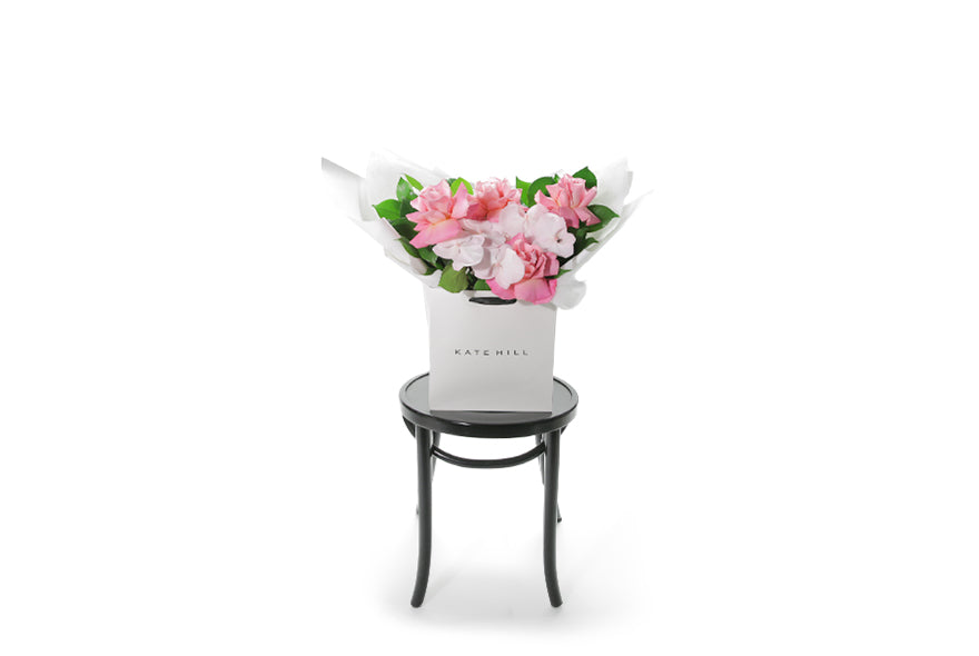 Best selling pastel pink seasonal flower bouquet displaying mixed pink flowers and green foliage. Large bouquet presented in Kate Hill Flower Bag. Large bouquet bag sitting on a black bentwood chair. Wide image to show the size of the bouquet.