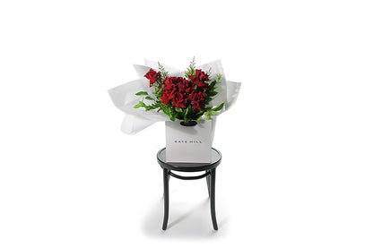 Classic and large red rose bouquet displaying 12 stems of red reflexed roses and seasonal green foliage. Valentines day or Romance gift bouquet presented in our Kate Hill Flower Bag. DIOR boasts a collection of short to medium-length red roses intertwined with seasonal foliage. Bouquet Bag sitting on a black bentwood chair. Wide image of the bouquet bag sitting on a black bentwood chair.