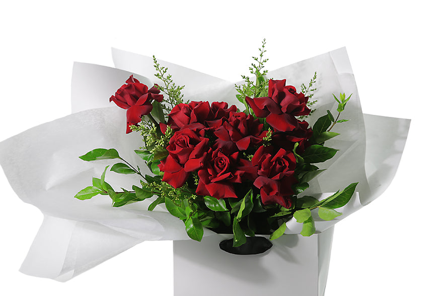 Classic and large red rose bouquet displaying 12 stems of red reflexed roses and seasonal green foliage. Valentines day or Romance gift bouquet presented in our Kate Hill Flower Bag. DIOR boasts a collection of short to medium-length red roses intertwined with seasonal foliage. Bouquet Bag sitting on a black bentwood chair. Close up image of the bouquet bag.