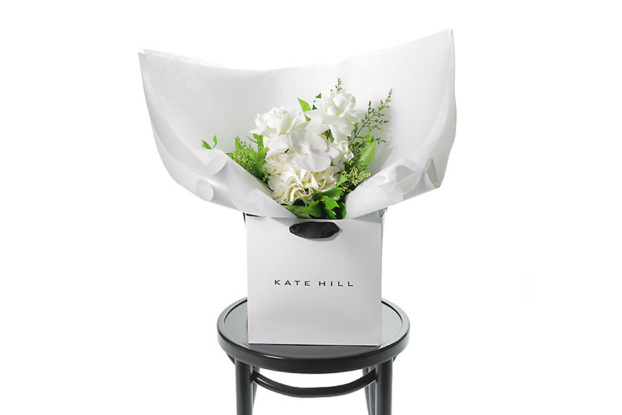 ELLA white and green posy wrapped in signature Kate Hill flowers wrapping and placed into a flower bag. Bouquet bag is sitting on a black bentwood chair. Seasonal white and green posy.