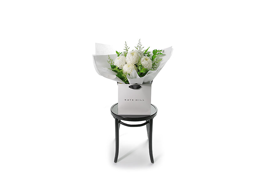 Wide image of the FLORENCE flower bouquet. FLORENCE is a hand tied bouquet crafted with 5 stems of white Disbuds and green seasonal foliages which are beautifully gift wrapped in our signature wrapping. Bouquet is placed into a Kate Hill flower bag. Flower flower bag is sitting on a black bentwood chair.