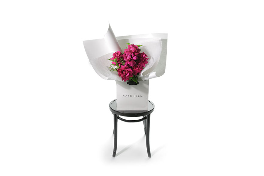 Wide image of the Floyd Rose Bouquet. Best selling hot pink rose bouquet named Floyd featuring 6 stems of hot pink reflexed roses and green seasonal foliage. Gift bouquet presented in our Kate Hill Flower Bag. Floyd rose bouquet is sitting on a black bentwood chair.