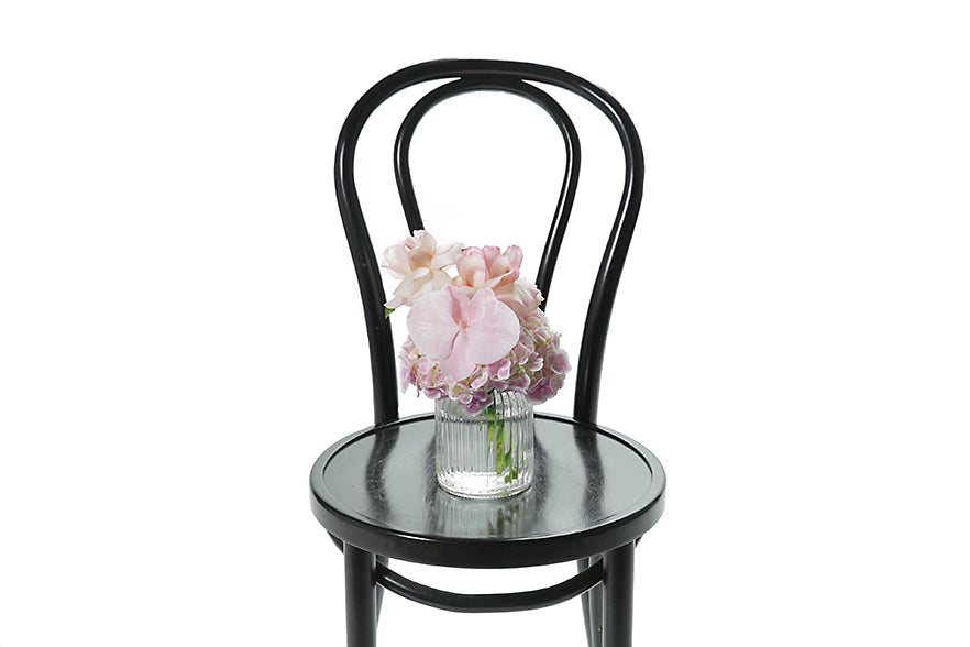 Petite ribbed glass vase displaying soft pastel pink seasonal flower. Gift includes the vase. Petite pastel vase is sitting on a black bentwood chair.