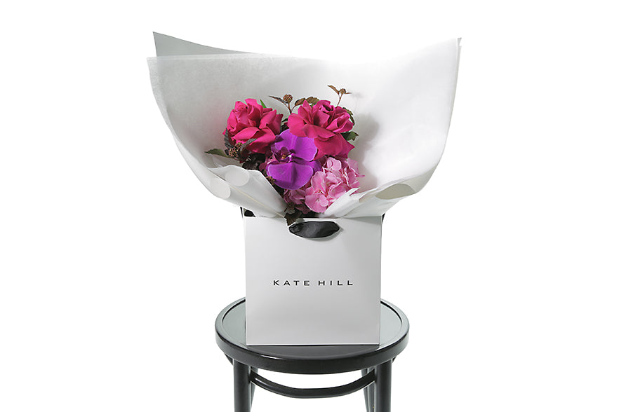 Small bright pink and magenta pink posy bouquet. Pink flowers and green seasonal foliage. Presented in Kate Hill Flower Bag. Bouquet bag is sitting on a black bentwood chair.