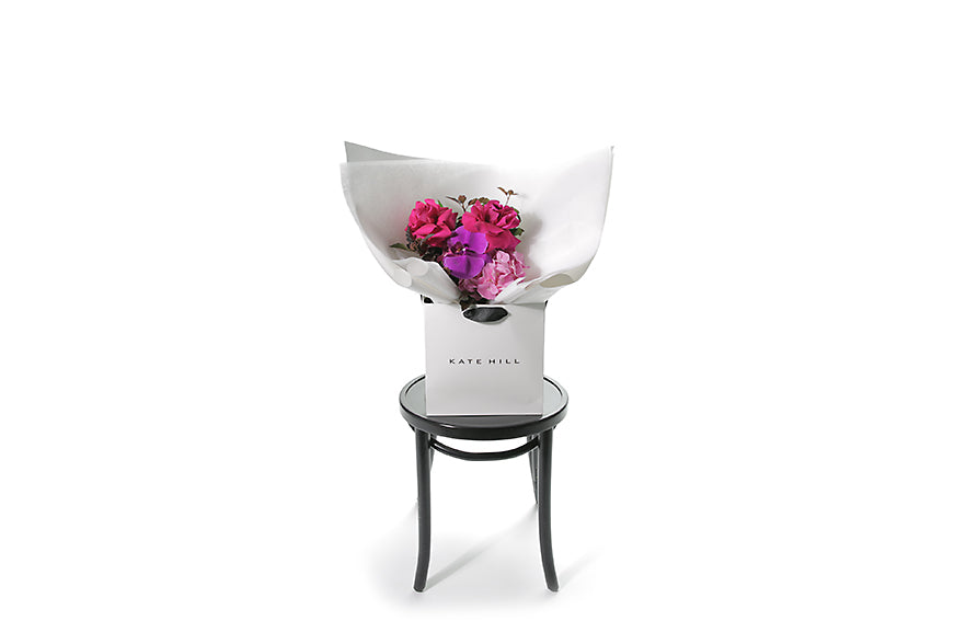 Wide image of the LENI flower bouquet. Petite in size and vibrant in colour. Small bright pink and magenta pink posy bouquet. Pink flowers and green seasonal foliage. Presented in Kate Hill Flower Bag. Bouquet bag is sitting on a black bentwood chair.