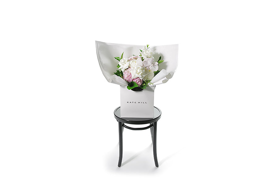 Pastel pink and white flower bouquet with seasonal foliage. Gift Bouquet presented in Kate Hill Flower Bag. Medium sized bouquet sitting on a black bentwood chair with white background. Wide image of the bouquet.