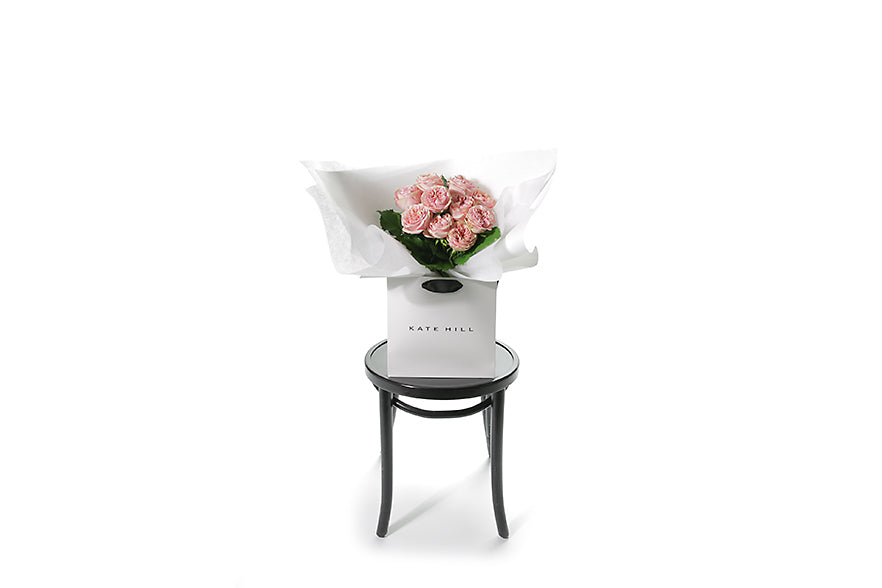 Wide image of the VERA rose flower bouquet. VERA is classed as a small sized, hand tied bouquet of 10 stems of Classic Roses (no additional foliage with roses only). Vera rose bouquet is beautifully wrapped in our signature wrapping and placed into our flower bag. Bouquet bag is sitting on a black bentwood chair.