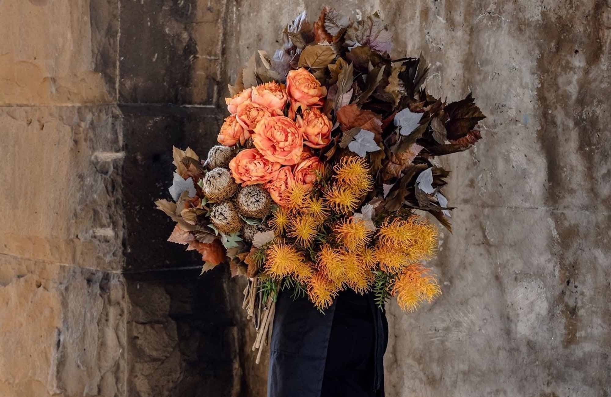Melbourne florist holding large bunches of artificial flowers in Orange and brown hues against backdrop of wall