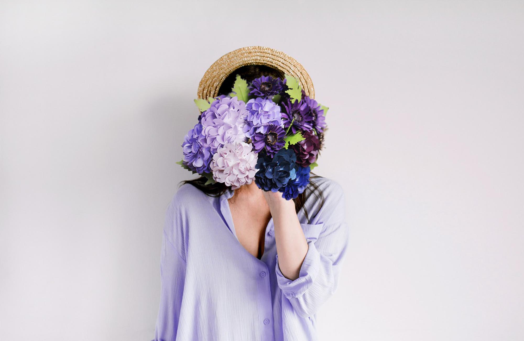 Lady in purple shirt with straw hat holding purple hydrangeas in front of her face with a grey background
