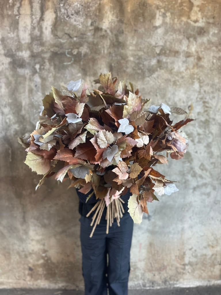 Florist wearing black holding a big bunch of Artificial stems of faux maple foliage, standing in front of a concrete wall.