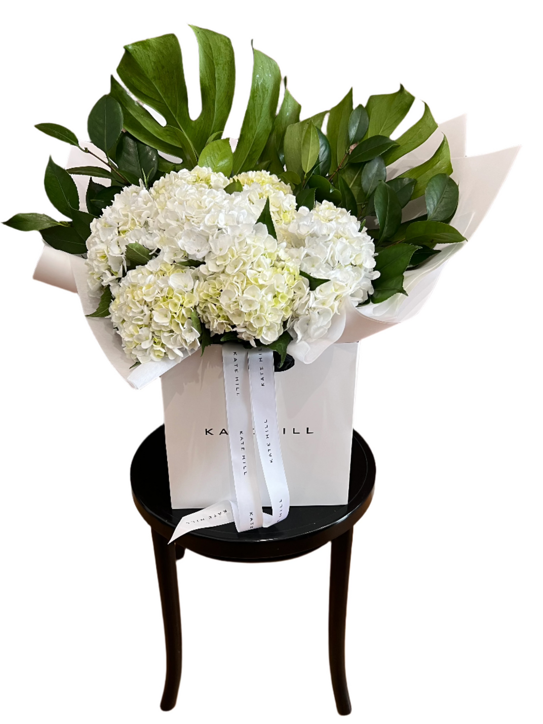 A large bouquet of white hydrangeas and green foliages wrapped beautifully in our Kate Hill Flower bag which is sitting on a black bentwood chair and white background.