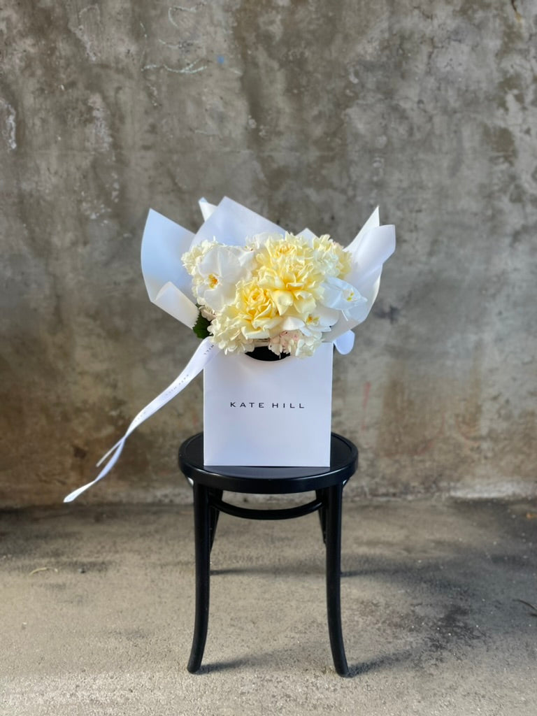 A lemon and white bouquet beautifully wrapped in our signature presentation, bouquet displayed in our flower bag sitting on a black bentwood chair in front of a concrete wall.