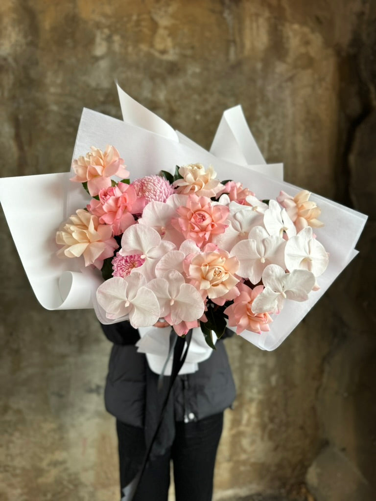 Close up image of blush MILA bouquet. A luxe blush flower bouquet wrapped in signature white wrapping. Florist in black holding the blush flower bouquet in front of a concrete wall.