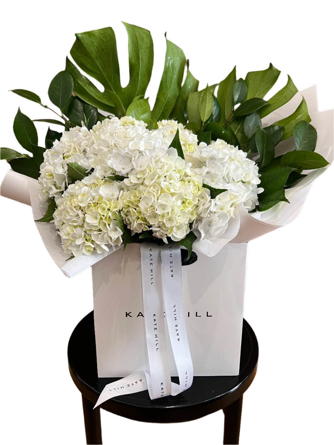 A close up of a large bouquet of white hydrangeas and green foliages wrapped beautifully in our Kate Hill Flower bag which is sitting on a black bentwood chair and white background.