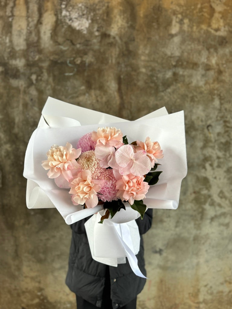 Wide image of the Chloe Blush Flower Bouquet. A close up of a blush pink flower bouquet, beautifully wrapped in white signature paper. Florist holding the bouquet in front of a concrete wall.