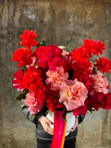 Florist holding the design. A large white vase displaying masses of orange, pink and watermelon roses which is standing against concrete wall.