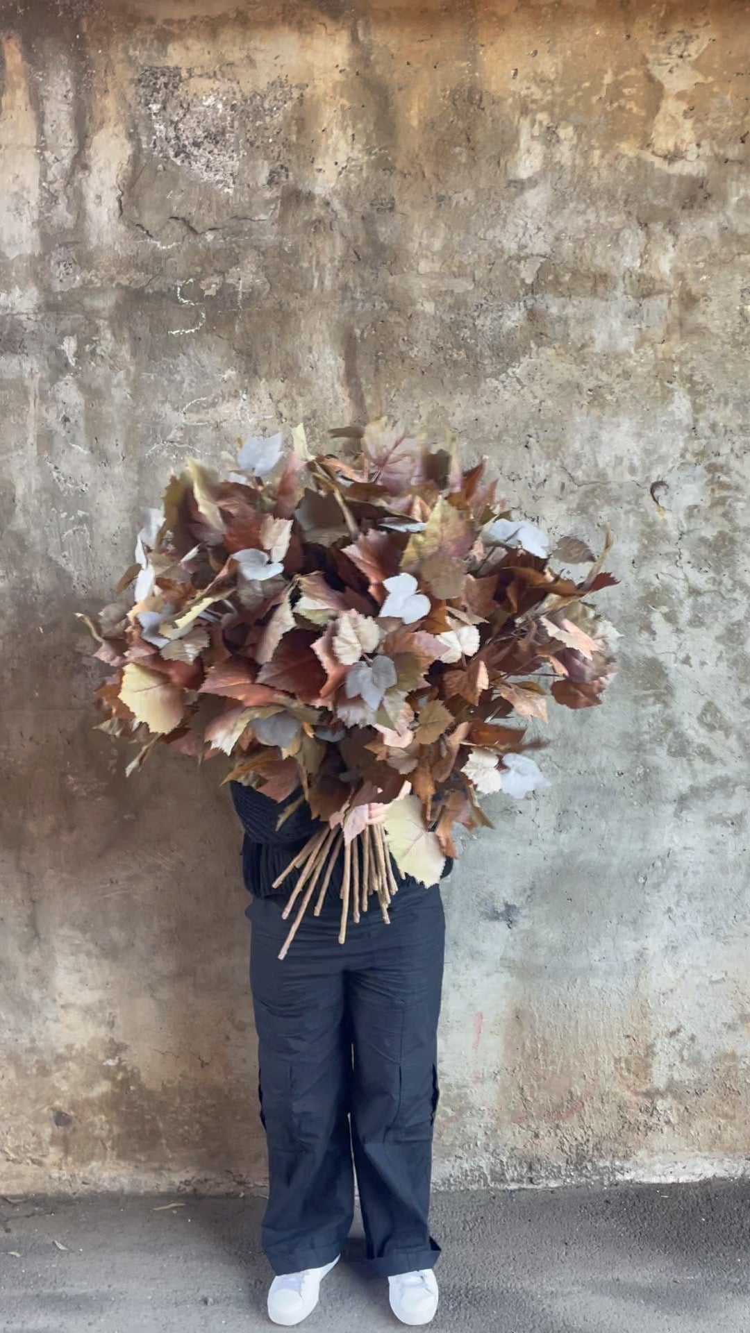 Video of Florist wearing black holding a big bunch of Artificial stems of faux maple foliage, standing in front of a concrete wall.