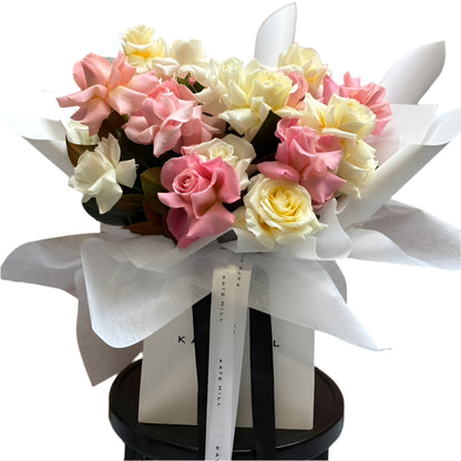 Bouquet of pastel mix roses in pastel pink, buttery lemon and white roses with a black and white box against a white wall.