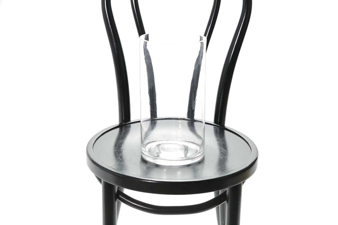 Close up image of 25cm clear glass vase. A 25cm clear glass vase sitting on a black bentwood chair with white wall background.
