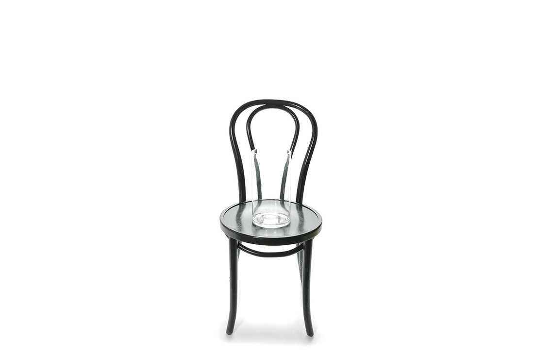 A 25cm clear glass vase sitting on a black bentwood chair with white wall background.
