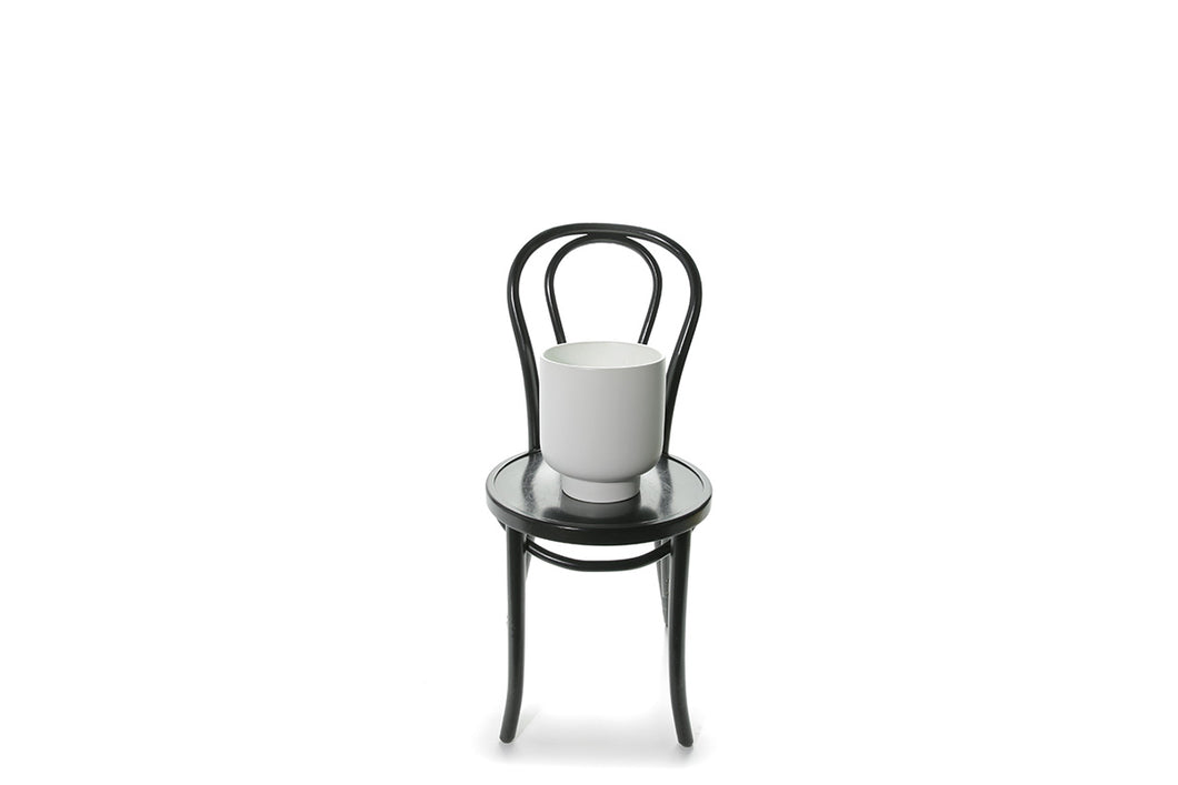 A large 25cm white ceramic vase sitting on a black bentwood chair with white backdrop.