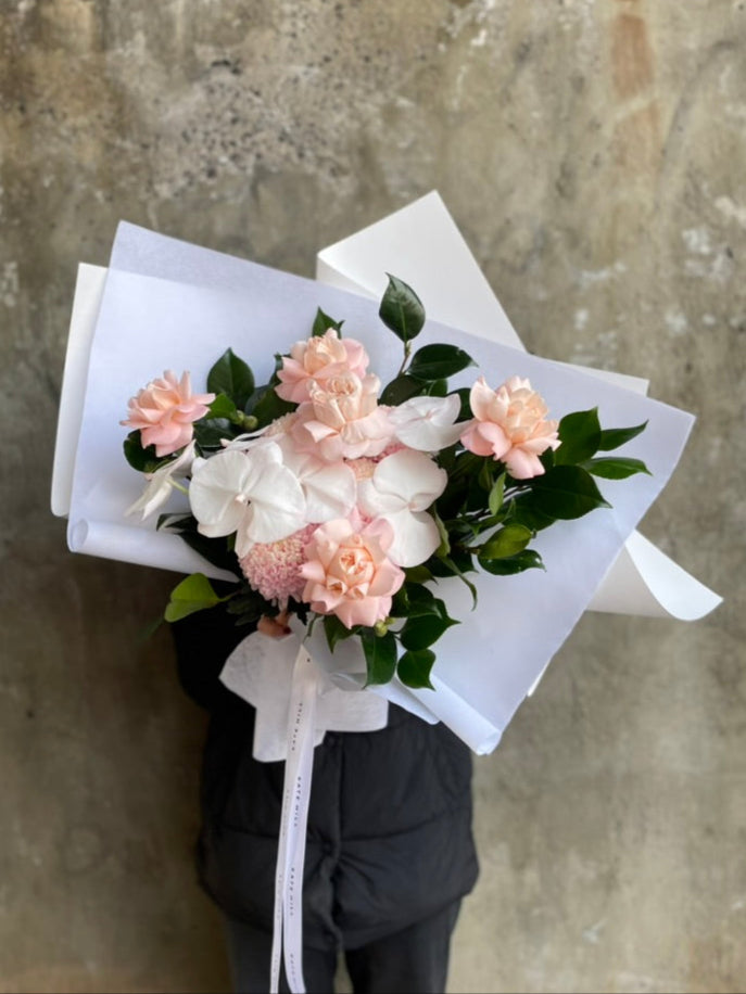 A blush flower bouquet held by a florist wearing black against a concrete grey wall.