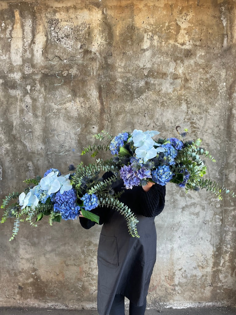 Up close image of florist holding 2 blue tablescapes. Florist holding a table hedge of blue seasonal flowers and grey blue foliages. Florist standing in front of a concrete wall.