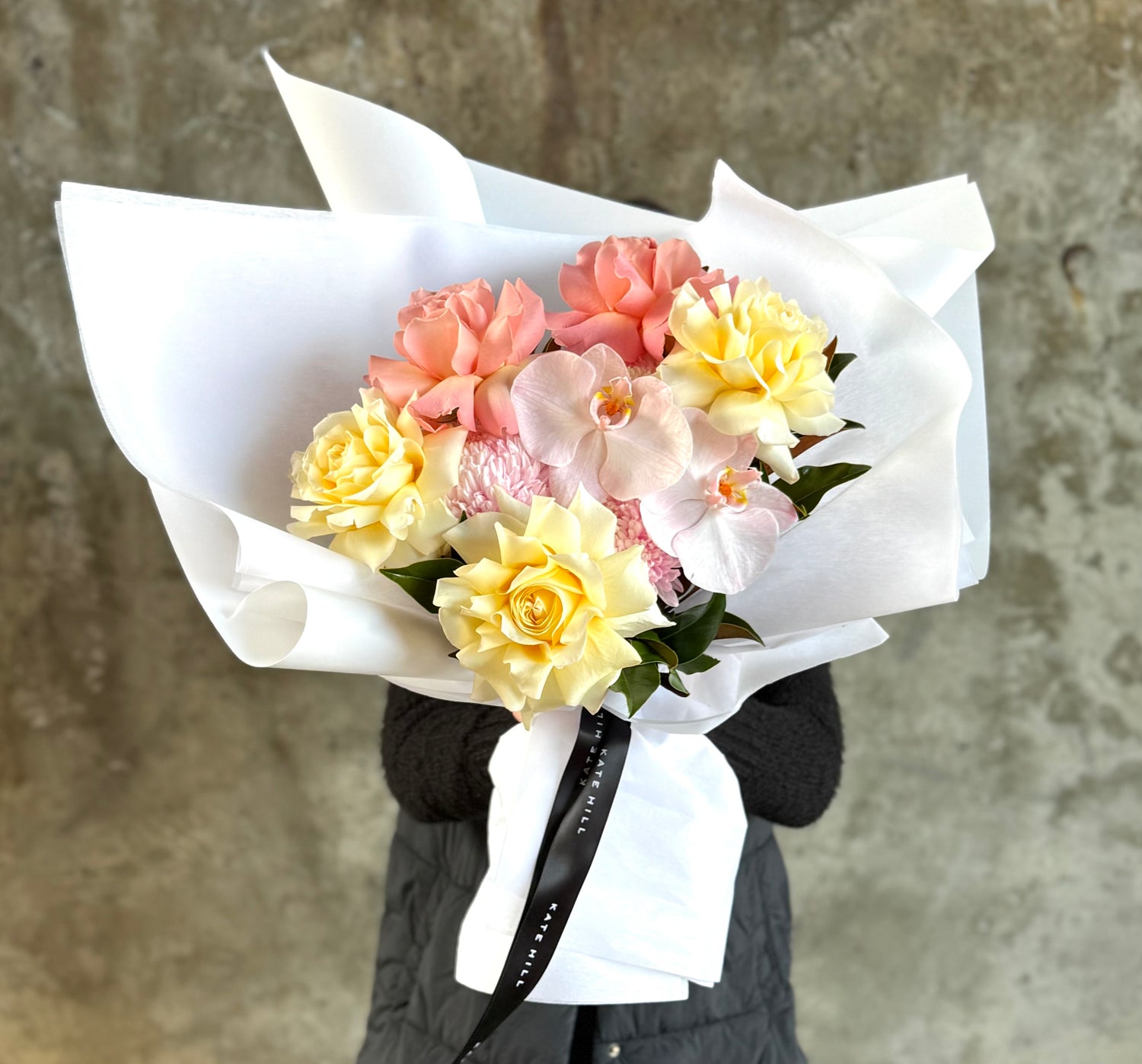 Close up image of florist holding a lemon and blush bouquet in front of a concrete wall.
