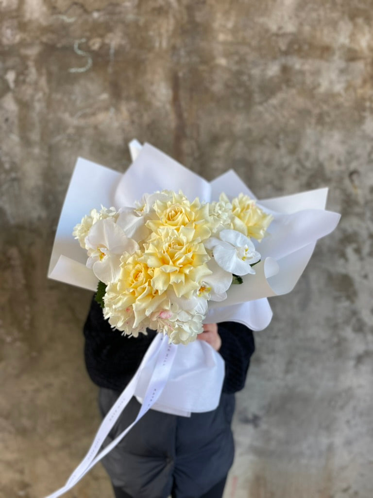 Florist holding our lemon and white bouquet beautifully wrapped in our signature presentation, bouquet displayed in our flower bag sitting on a black bentwood chair in front of a concrete wall.