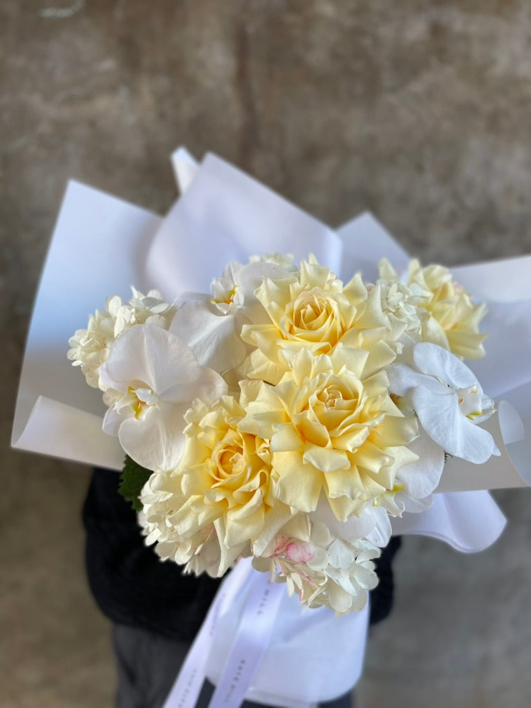 Close image of florist holding our lemon and white bouquet beautifully wrapped in our signature presentation, standing in front of front of a concrete wall.