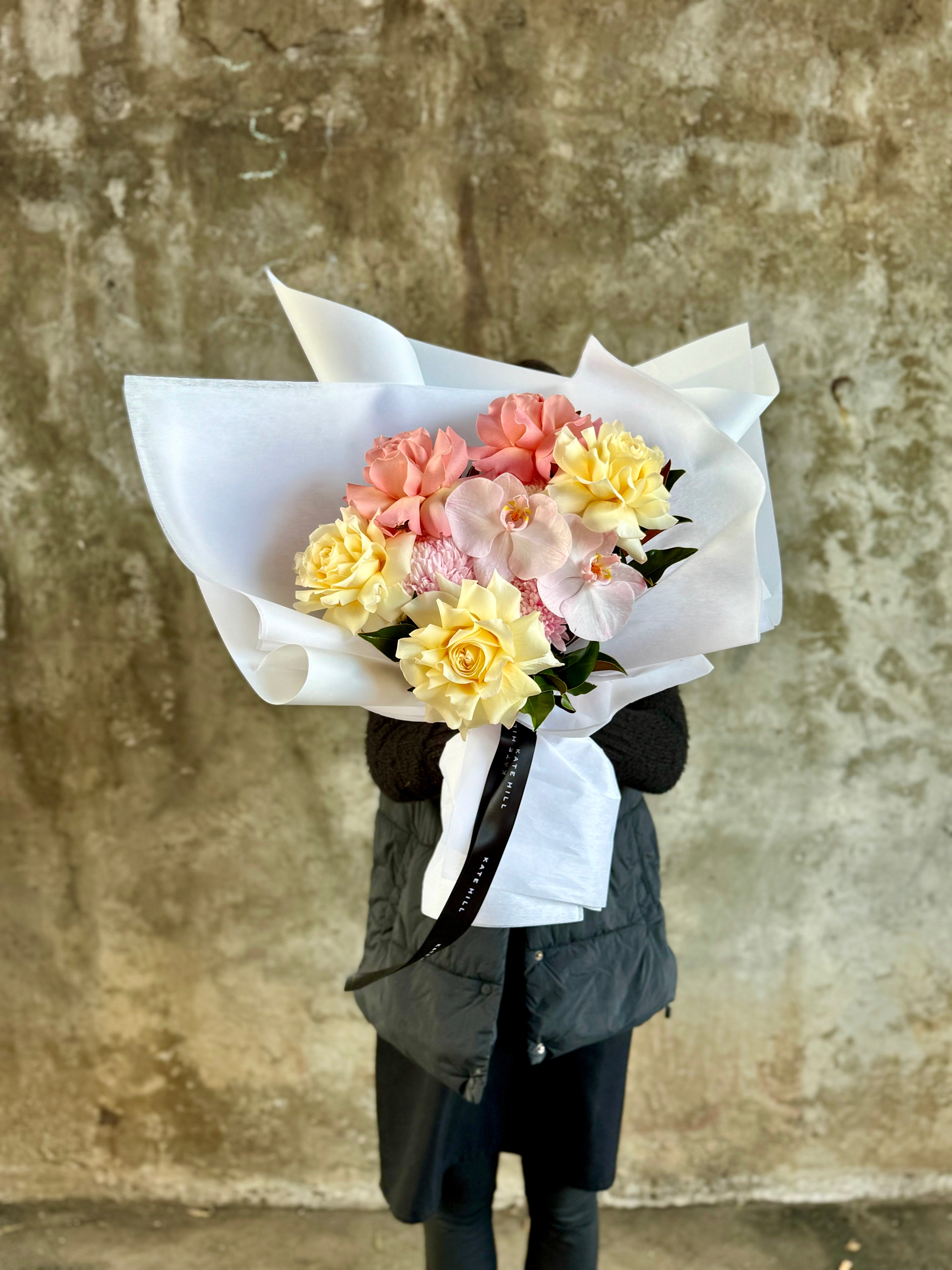 Florist wearing black clothing holding A medium sized lemon and blush guft bouquet in kate hill flower bag, sitting on a black bentwood chair in front of a concrete wall.