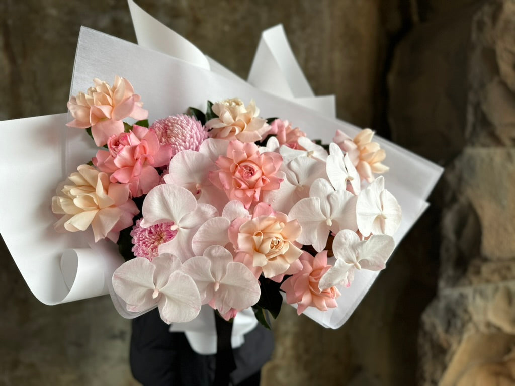 A luxe blush flower bouquet wrapped in signature white wrapping. Florist in black holding the blush flower bouquet in front of a concrete wall.