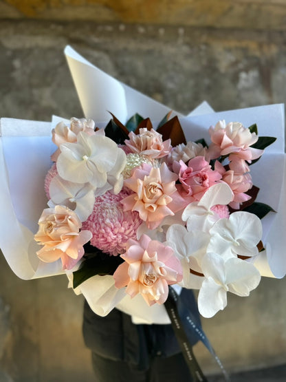 Close image of flower bouquet. A luxe blush flower bouquet wrapped in signature white wrapping. Florist in black holding the blush flower bouquet in front of a concrete wall.