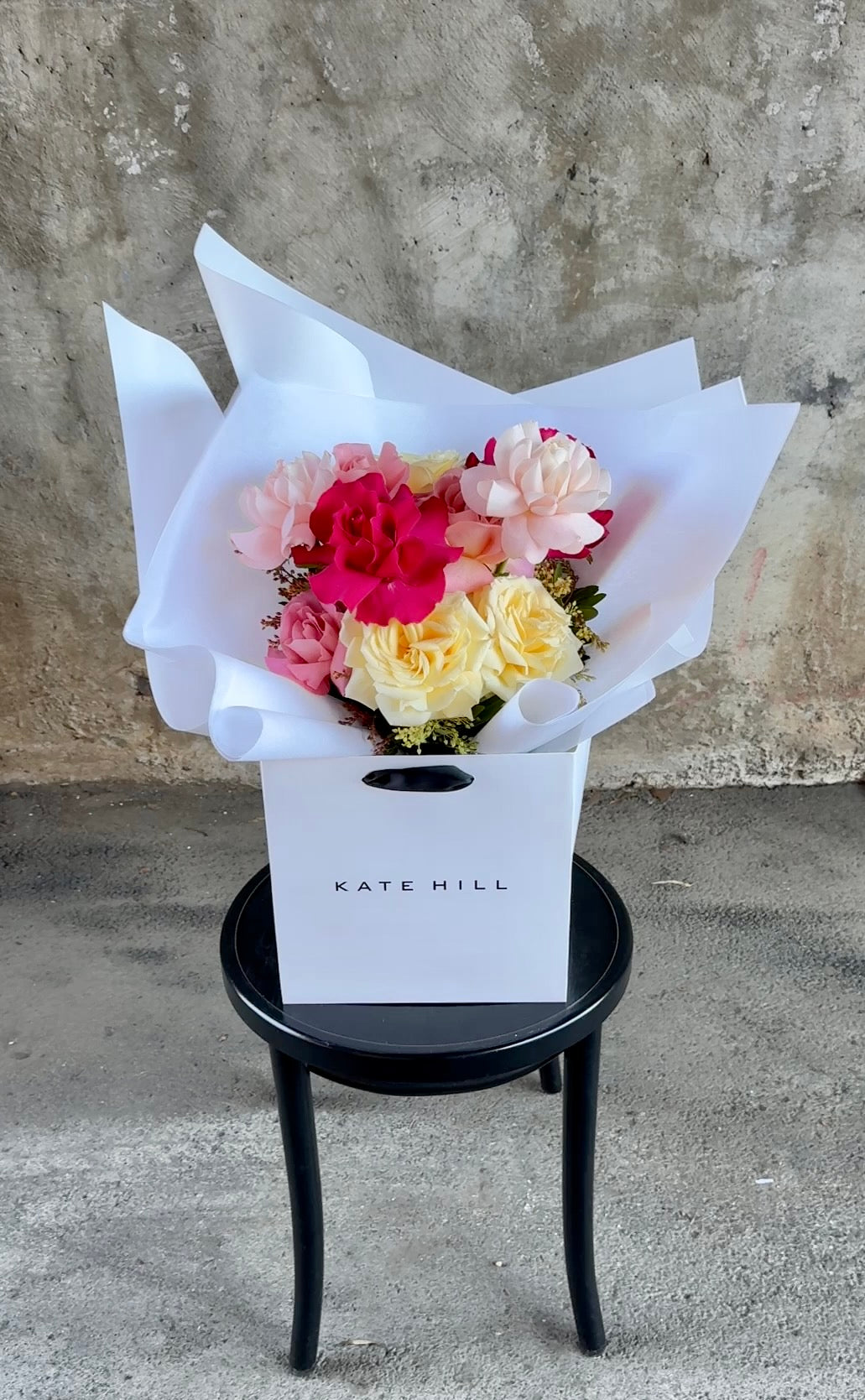 Video of mixed rose bouquet. A bouquet of mixed coloured reflexed roses, beautifully wrapped in white and placed into a KHF flower bag. Bouquet sitting on a black bentwood chair against a concrete wall.