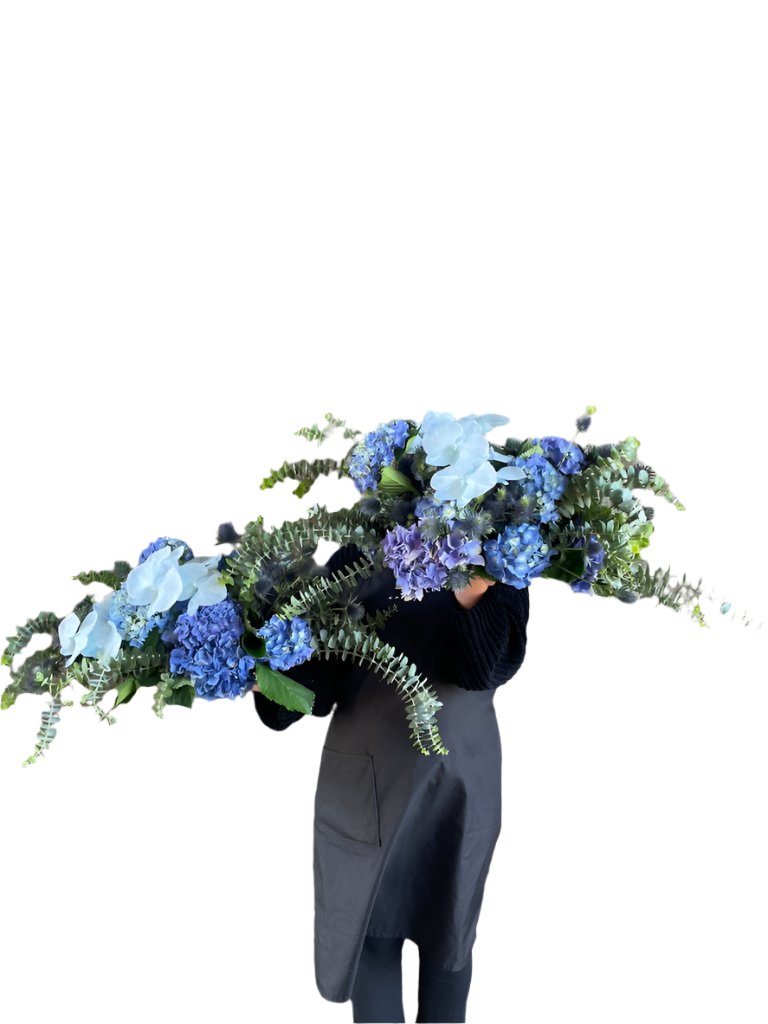 Florist holding 2 blue tablescape designs. Florist holding a table hedge of blue seasonal flowers and grey blue foliages. Florist standing in front of a concrete wall.