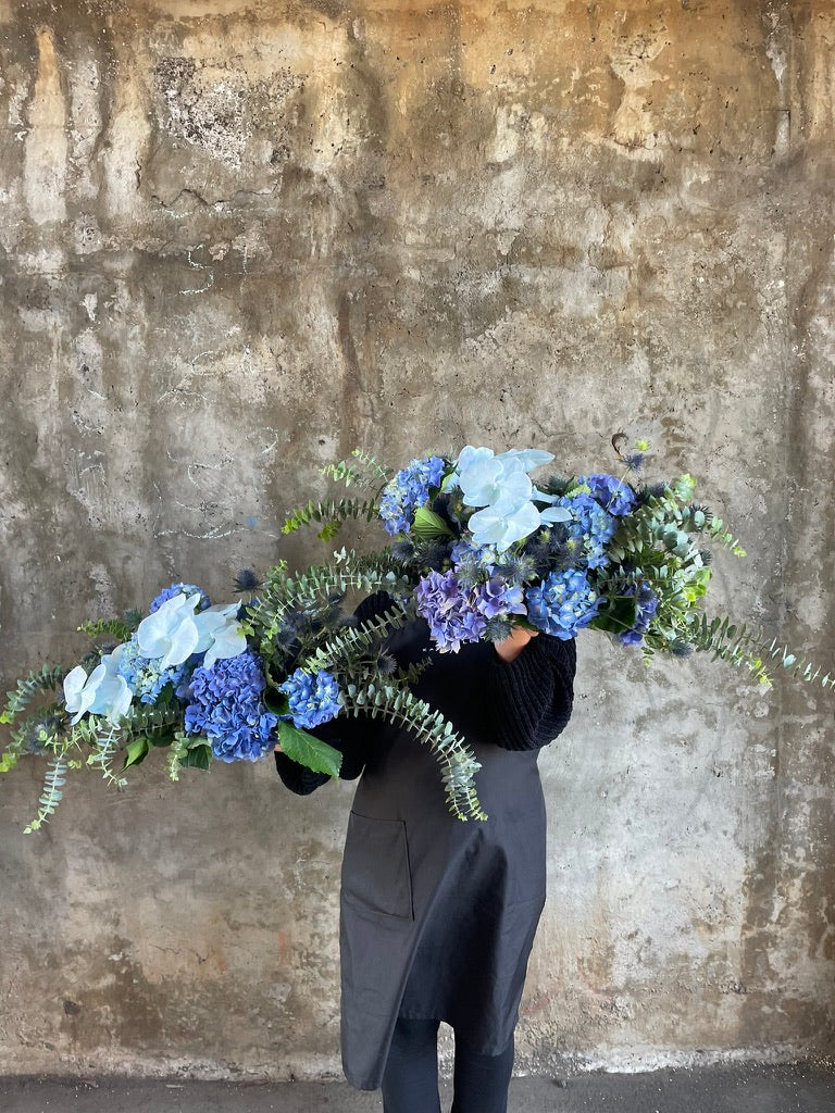 Florist holding 2 blue tablescape designs. Florist holding a table hedge of blue seasonal flowers and grey blue foliages. Florist standing in front of a concrete wall.