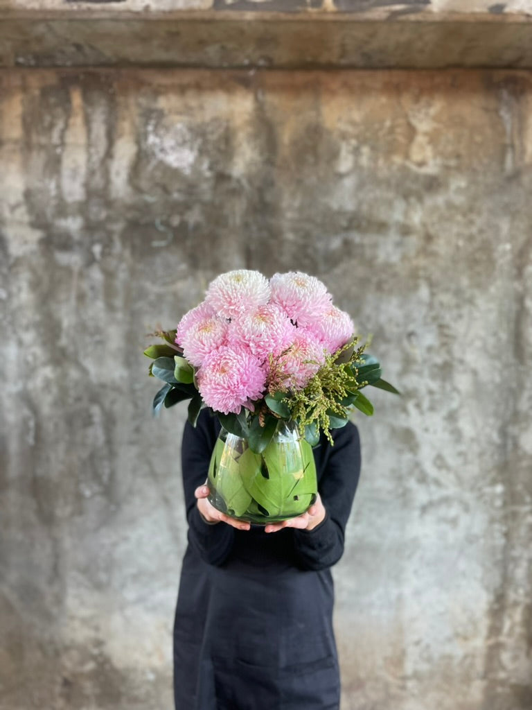 Florist holding A tapered vase displaying blush pink disbuds and foliage, in front of a concrete wall.