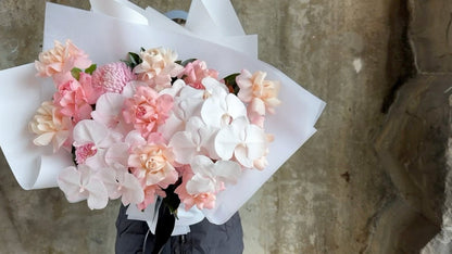 Video of luxe blush flower bouquet. A luxe blush flower bouquet wrapped in signature white wrapping. Florist in black holding the blush flower bouquet in front of a concrete wall.