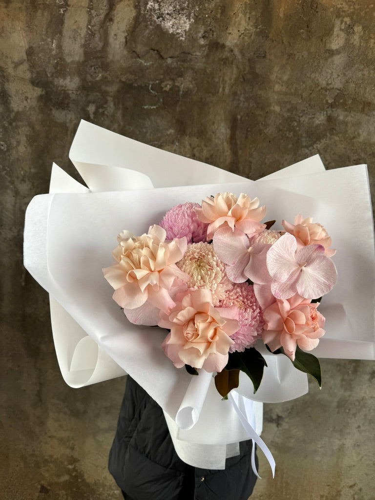 Side angle of florist holding gift bouquet. A blush birthday bouquet of blush disbud, blush roses and blush phalaenopsis which is beautifully wrapped in KHF signature style. Florist wearing black and holding the guft bouquet against a concrete wall. 