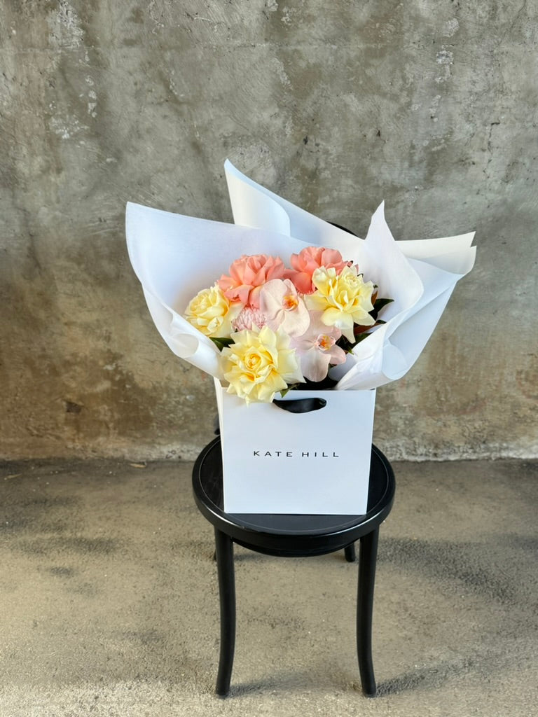 A medium sized lemon and blush guft bouquet in kate hill flower bag, sitting on a black bentwood chair in front of a concrete wall.
