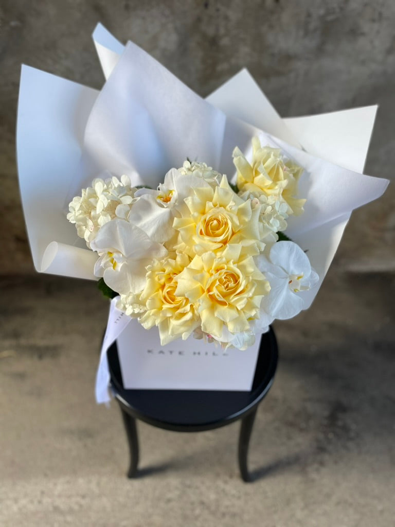 Image from above of a lemon and white bouquet beautifully wrapped in our signature presentation, bouquet displayed in our flower bag sitting on a black bentwood chair in front of a concrete wall.