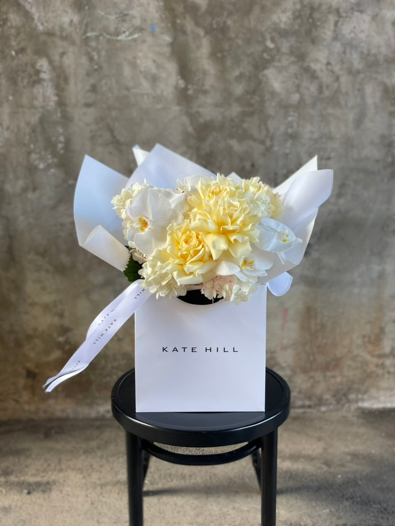 Close up image of A lemon and white bouquet beautifully wrapped in our signature presentation, bouquet displayed in our flower bag sitting on a black bentwood chair in front of a concrete wall.